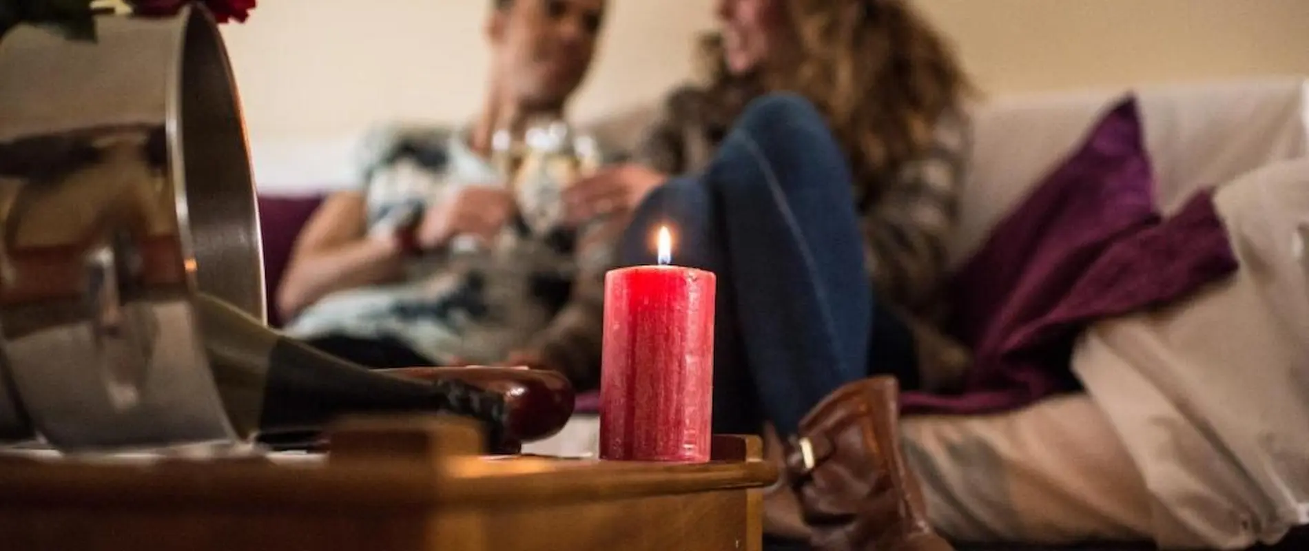 Pareja con copas de vino, vela roja encendida en primer plano, ambiente íntimo en la sala.