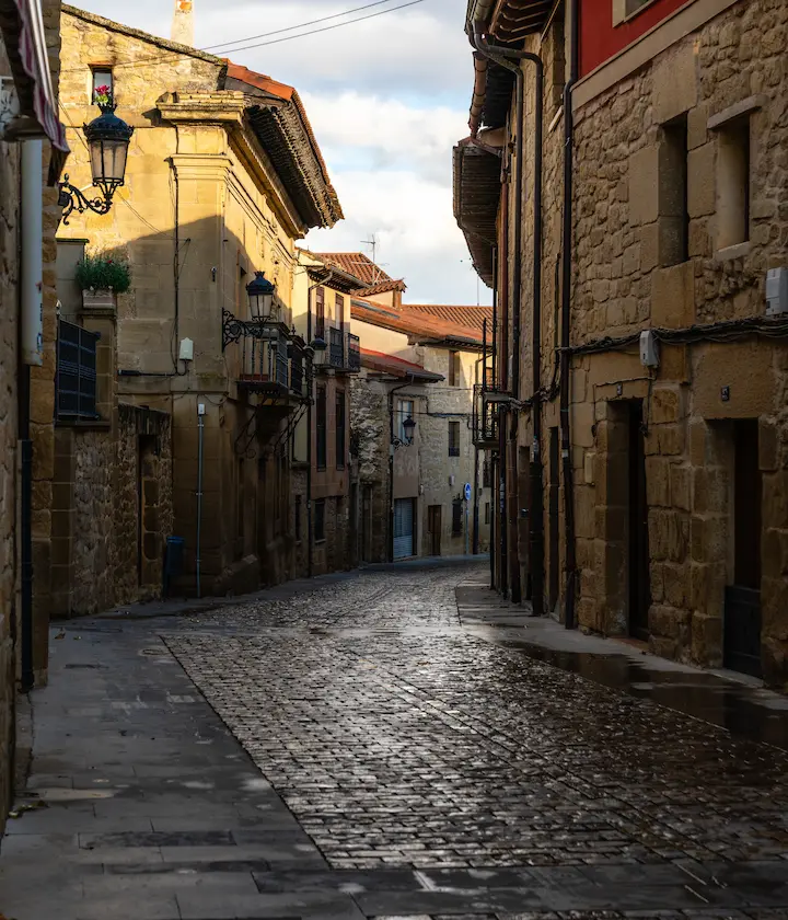 Calle estrecha empedrada entre edificios antiguos de piedra al atardecer.