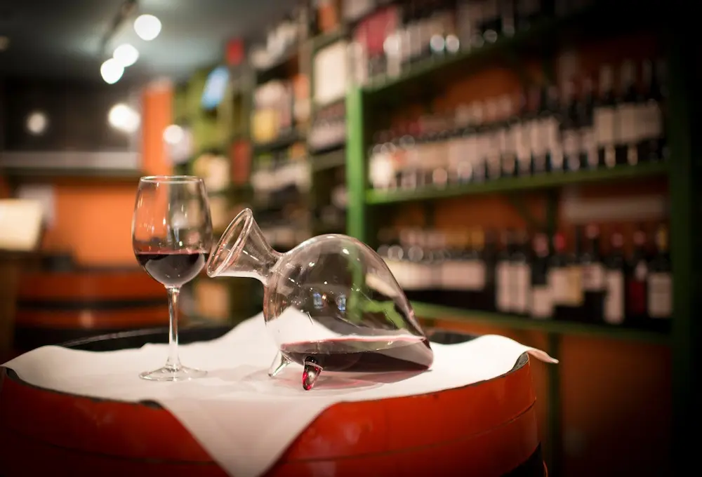 Una copa y un decantador de vino sobre un barril, en una bodega llena de botellas.