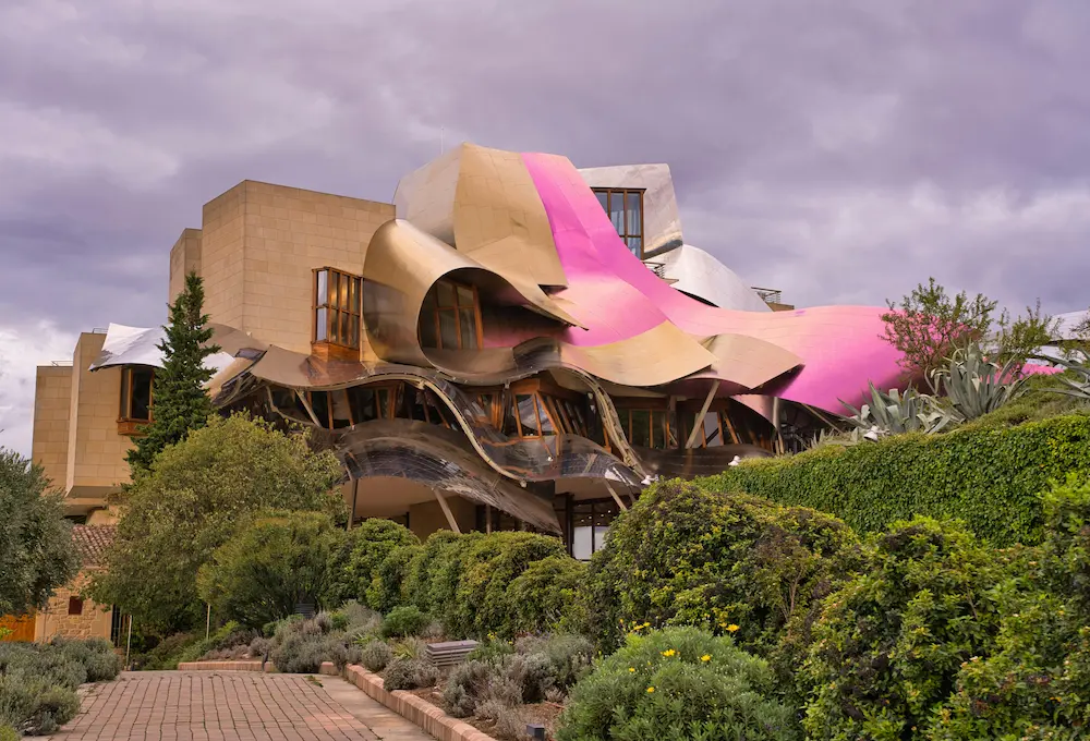 Edificio moderno con diseño ondulado y colores metalizados y rosa, rodeado de vegetación.