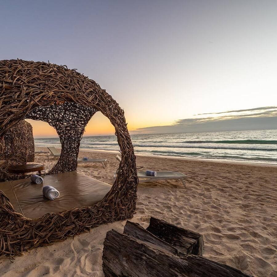 Wicker beach cabana at sunset, facing the ocean with lounge chairs and towels on the sandy shore.