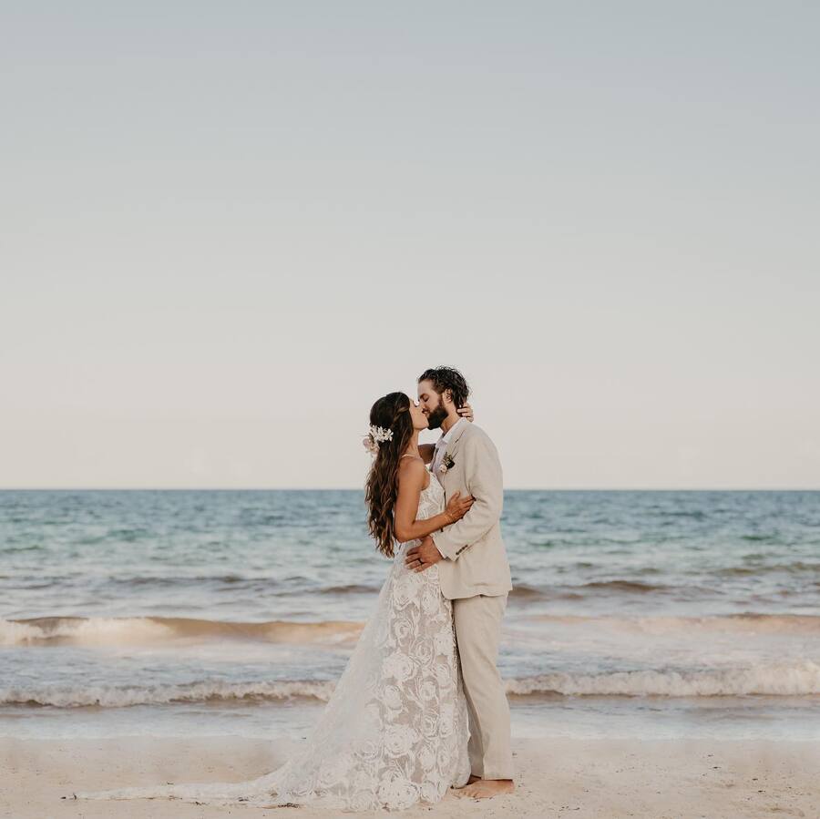 A couple embraces and kisses on a beach in wedding attire, with the ocean in the background.