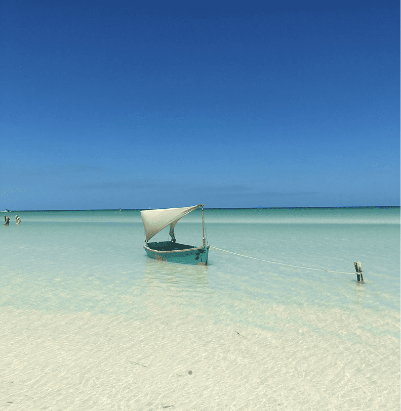 A small sailboat moored in clear, shallow water on a sunny day with a deep blue sky.