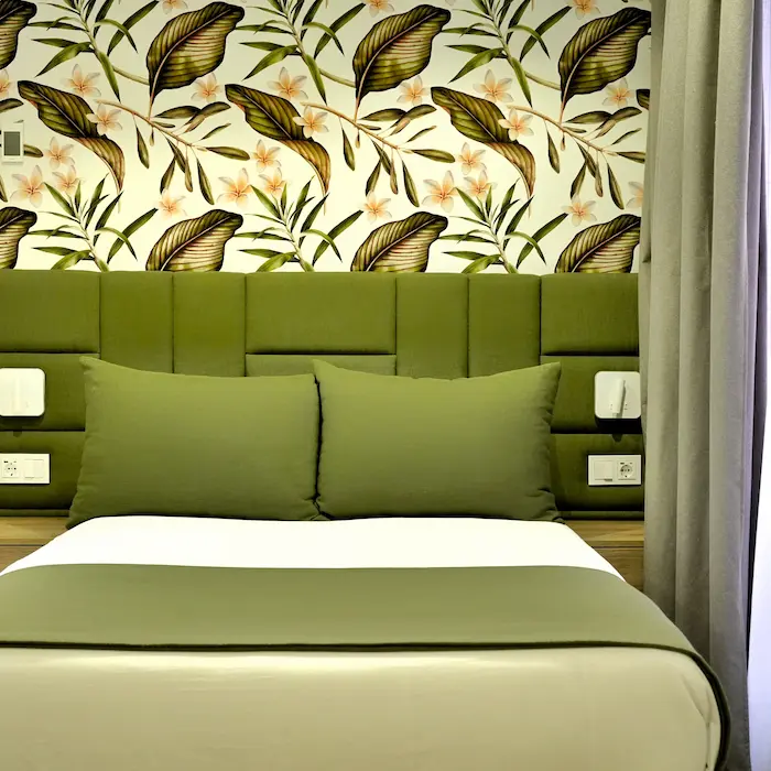 Cama con cojines verdes frente a papel tapiz de hojas y flores.