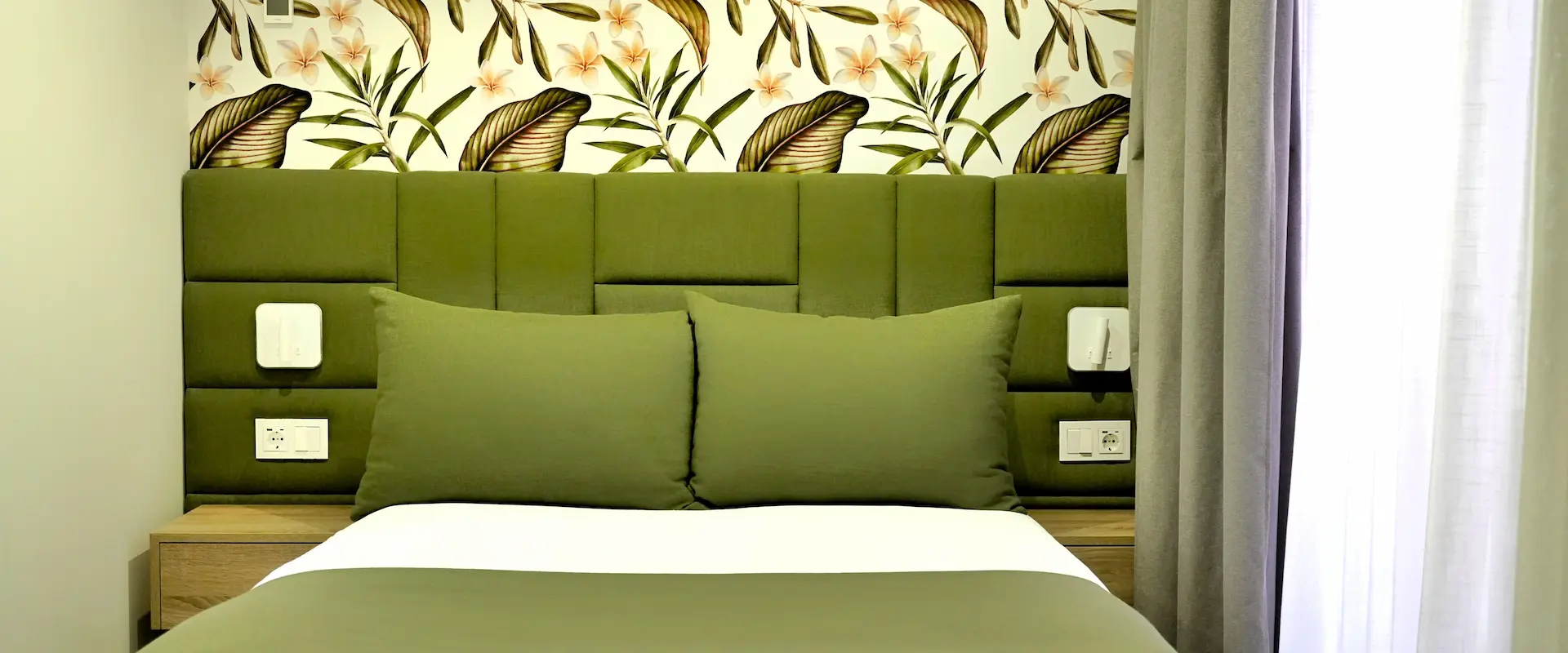 Cama con cabecera verde y mural floral, junto a cortinas y apliques en una habitación.