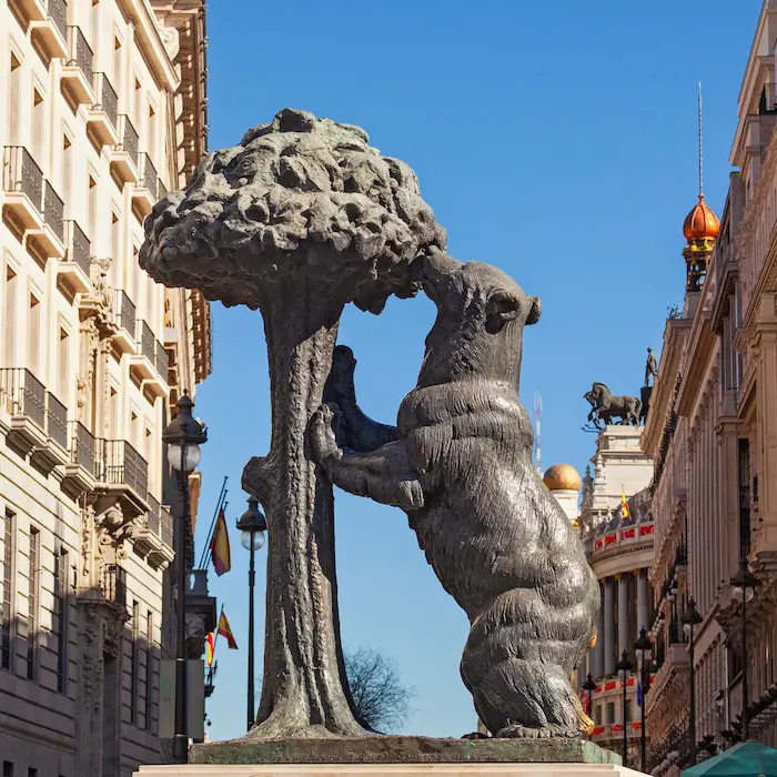 Estatua de un oso y un árbol en una plaza soleada con edificios alrededor.