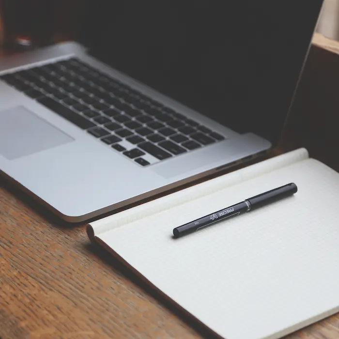 Laptop abierta junto a una libreta y un bolígrafo negro sobre una mesa de madera.