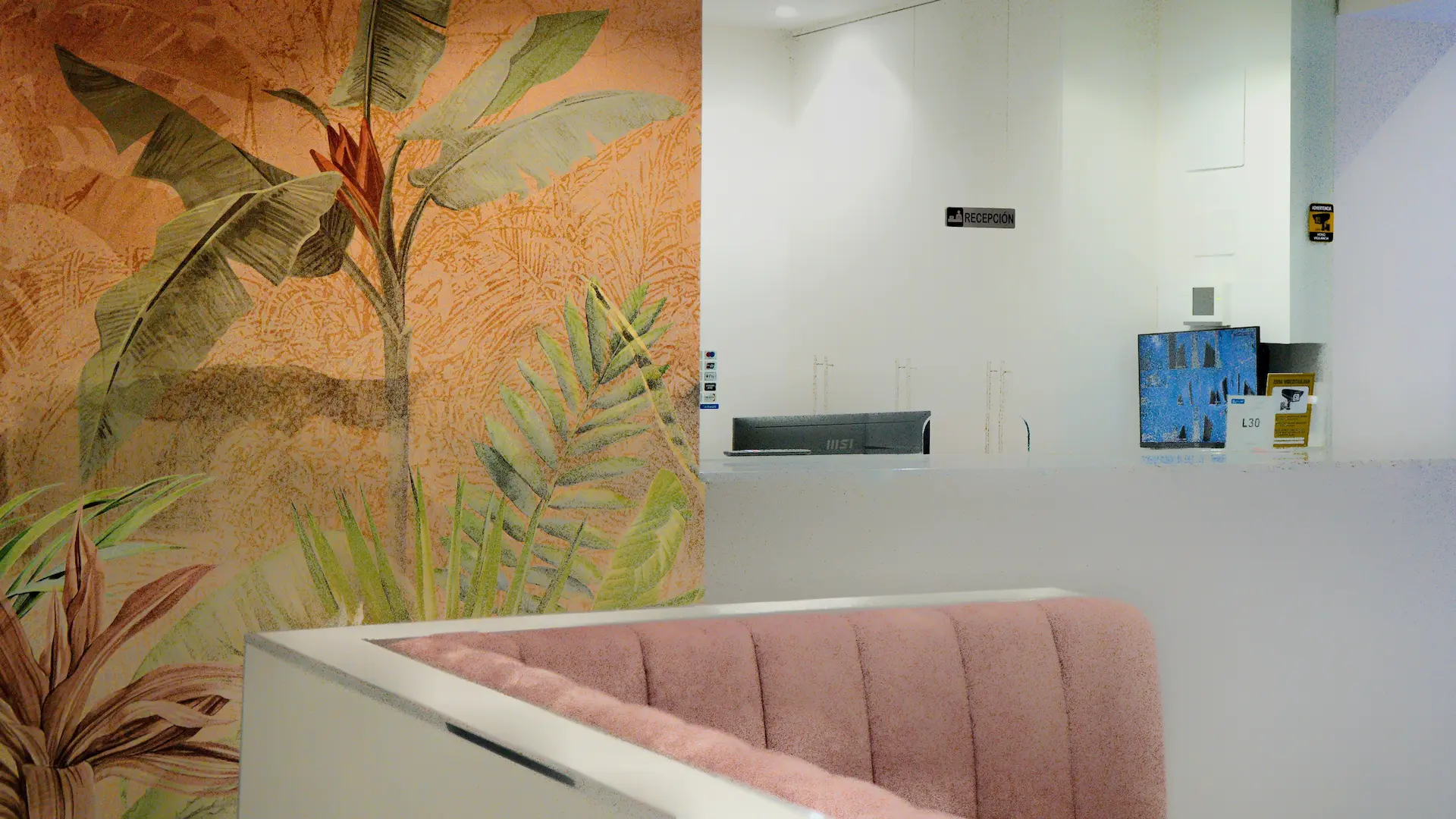 Pared decorada con hojas tropicales y mostrador minimalista de recepción junto a un sofá rosa.