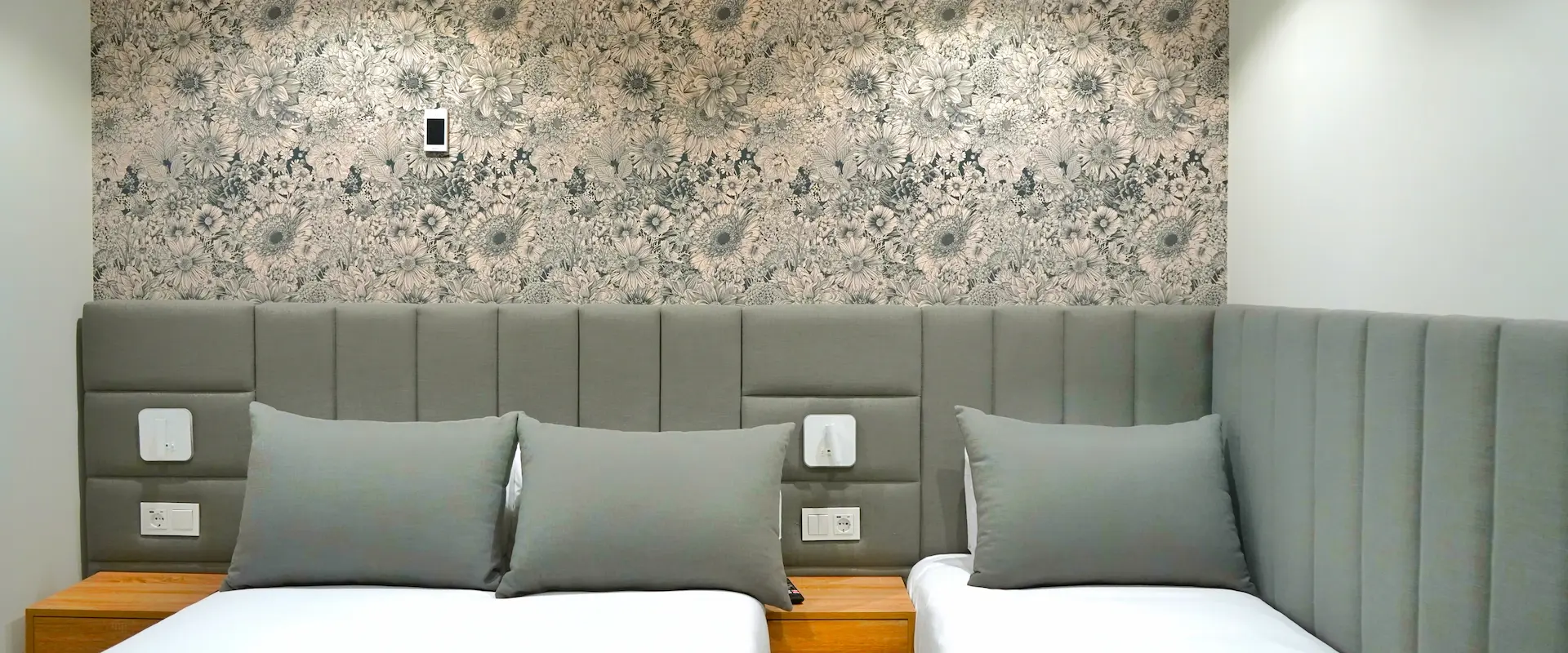 Habitación con dos camas, cojines grises y fondo de papel floral en tonos neutros.
