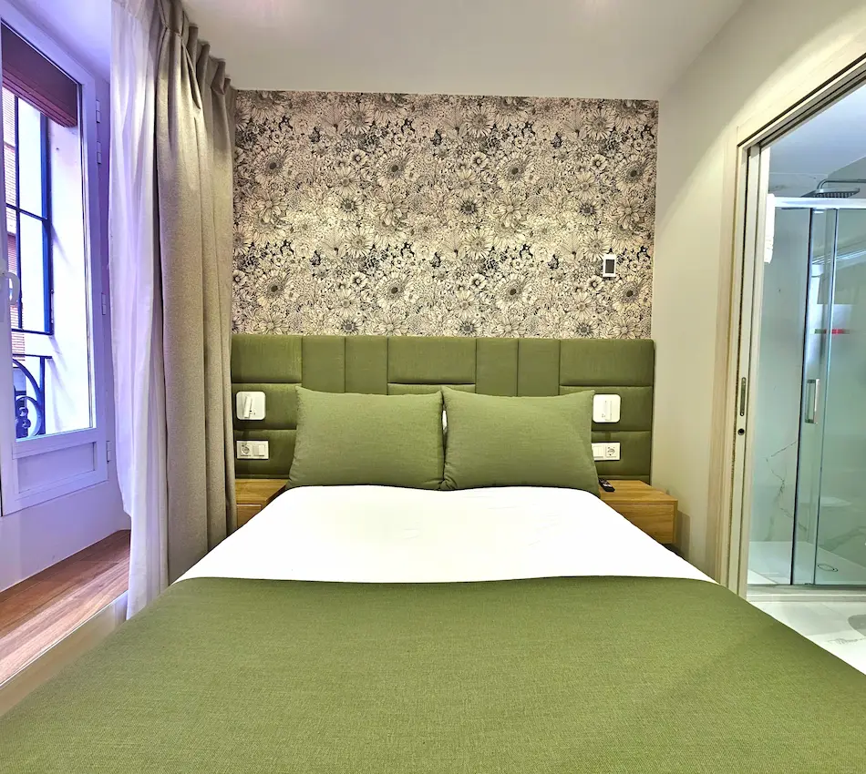 Cama con cubrecama verde y pared estampada en habitación bien iluminada junto a ventana y baño.