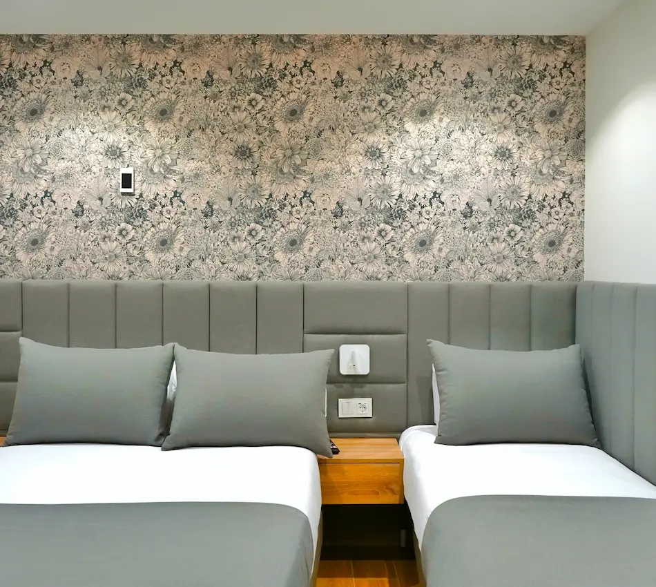 Dos camas individuales con cabeceras grises, fondo de papel tapiz floral y mesa de noche de madera.