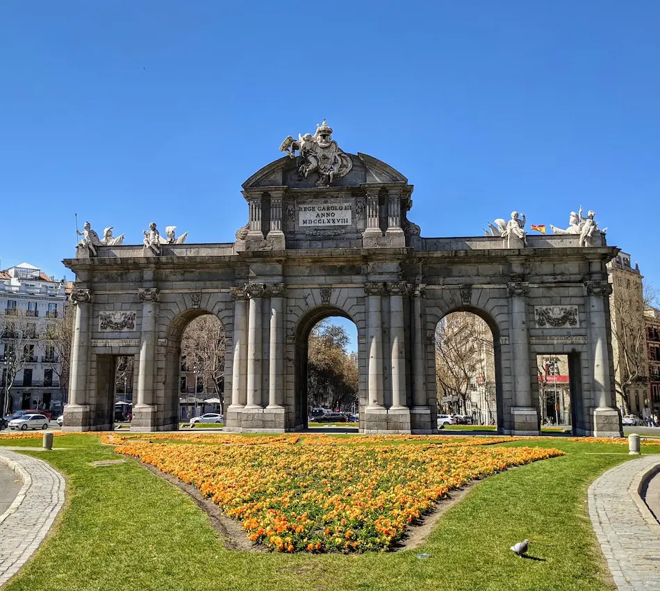 Arco de piedra con esculturas en plaza ajardinada, cielo azul despejado.