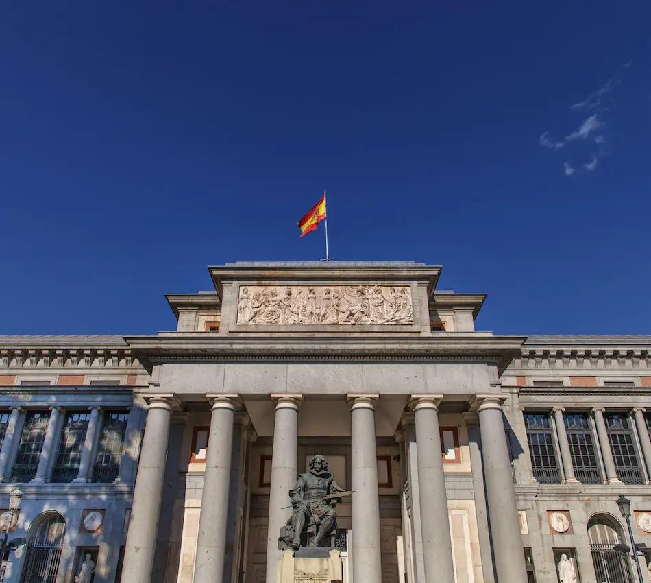 Edificio con columnas, estatua y bandera de España sobre un cielo azul.