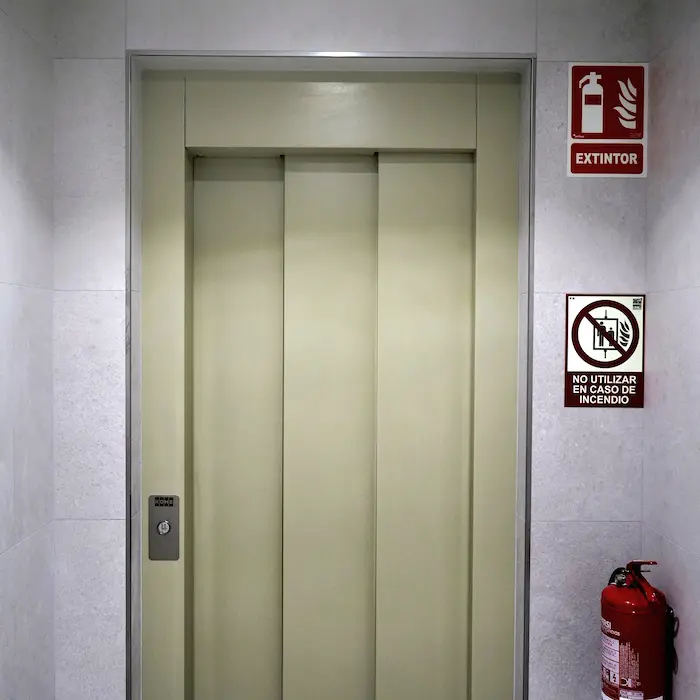 Puerta de un elevador con señales de no usar en incendio y un extintor rojo al lado.