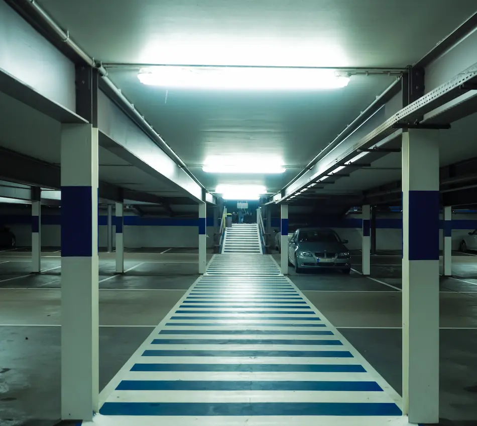 Estacionamiento subterráneo con columnas blancas y franjas azules, iluminado con luces fluorescentes.