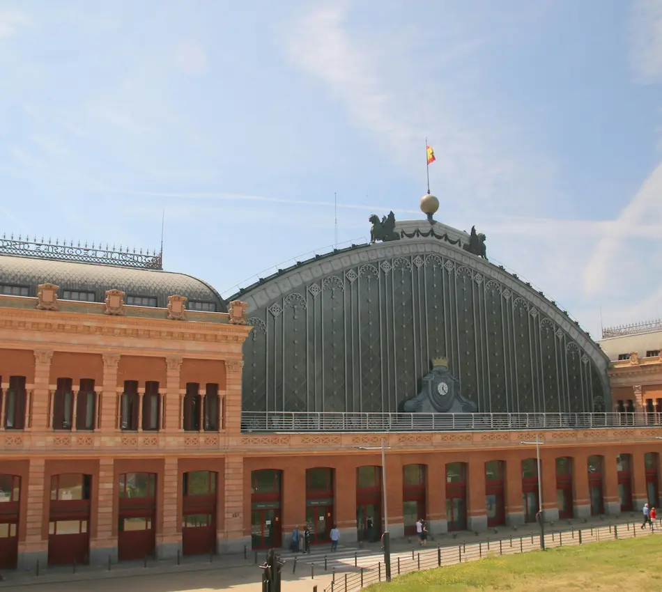 Estación de tren con fachada de ladrillo y techo arqueado, bandera ondeando arriba.