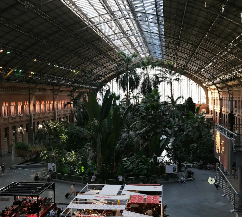 Estación cubierta llena de plantas tropicales, con puestos de comida y amplios ventanales iluminados.