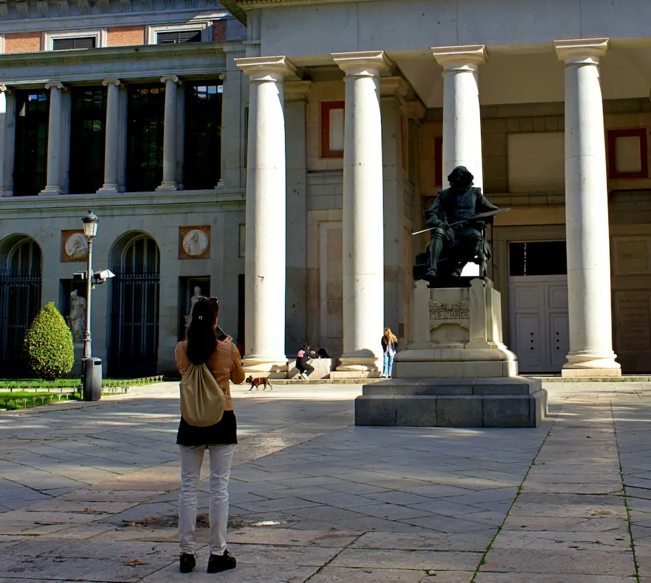 Persona toma foto de escultura frente a edificio con columnas en un día soleado.