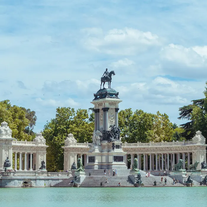 Monumento con estatua ecuestre frente a un lago, rodeado de árboles y cielo parcialmente nublado.