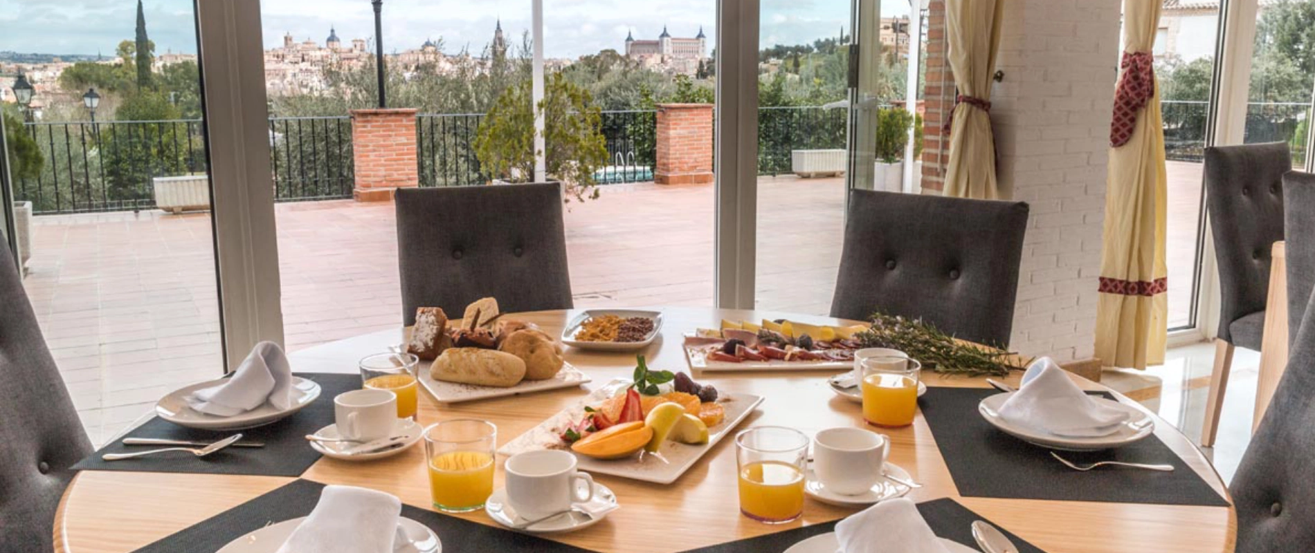 Mesa con desayuno variado: pan, frutas y jugo. Vistas panorámicas de una ciudad desde la ventana.