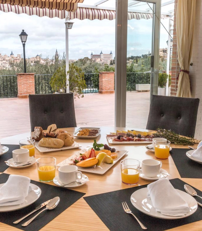 Mesa con desayuno servido, jugo, pan y frutas, con vista a la ciudad desde una terraza.