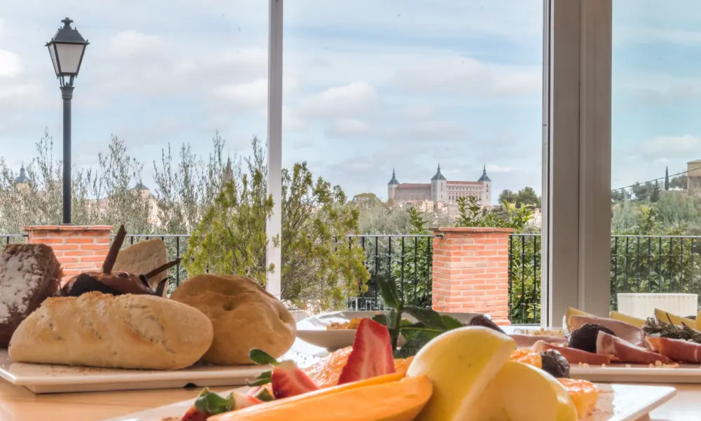 Mesa con panes y frutas, vista al exterior con un edificio antiguo a lo lejos.