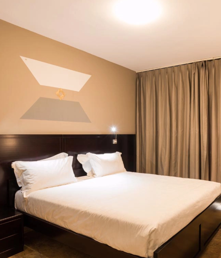 Habitación con cama doble, sábanas blancas y cortinas beige, iluminada cálidamente.