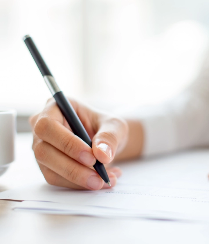 Una mano escribiendo con bolígrafo en papel, taza al fondo.