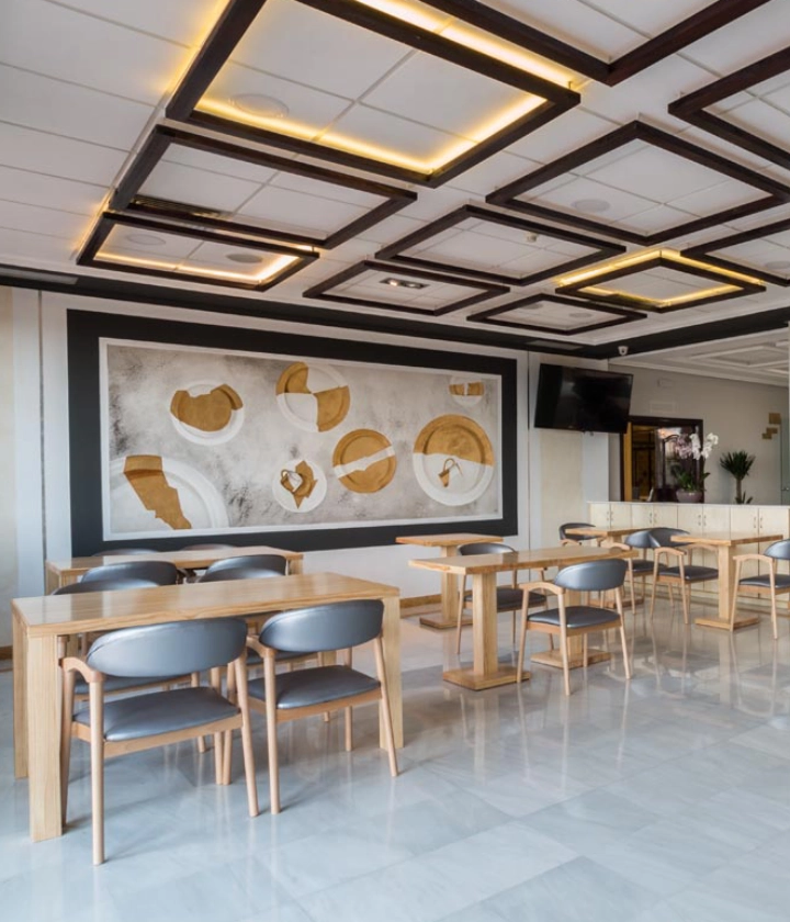 Sala moderna con mesas, sillas de madera, mural artístico y techo decorativo geométrico.