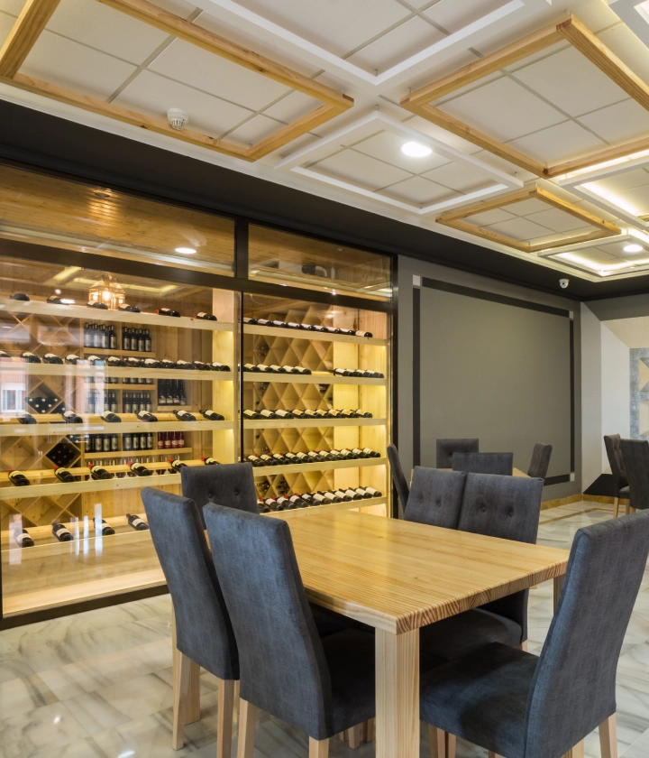 Mesa de madera con sillas grises frente a bodega con botellas de vino.