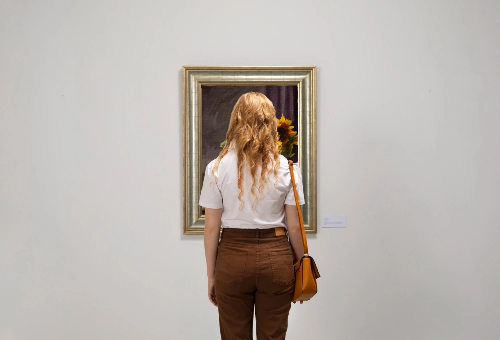 Mujer observando un cuadro en una galería de arte, de espaldas, con cabello rubio y bolso marrón.