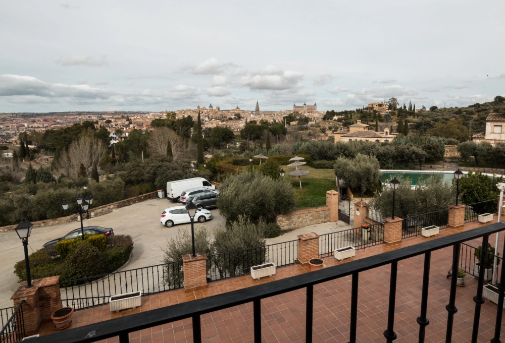 Vista de una ciudad desde una terraza con árboles, vehículos y una piscina en el paisaje.