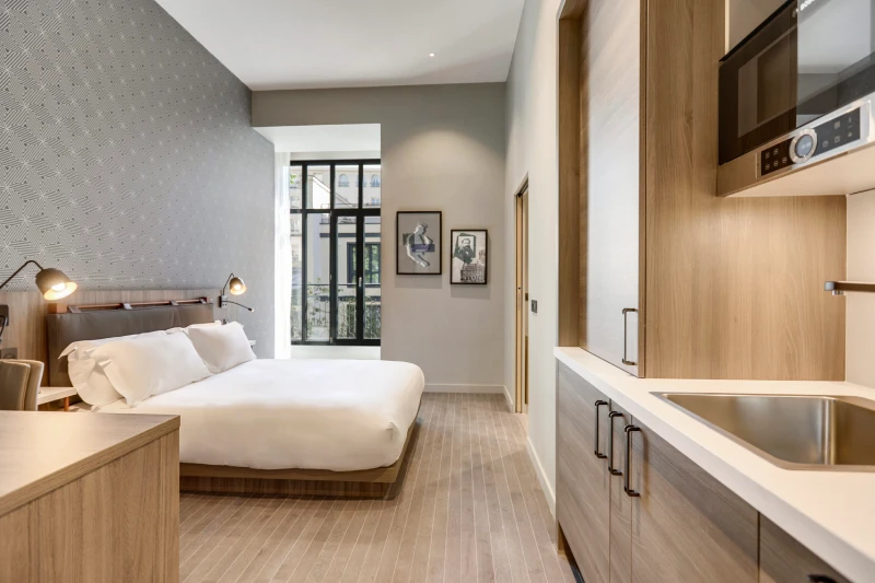 Chambre moderne avec un lit double, kitchenette et fenêtre donnant sur un balcon.
