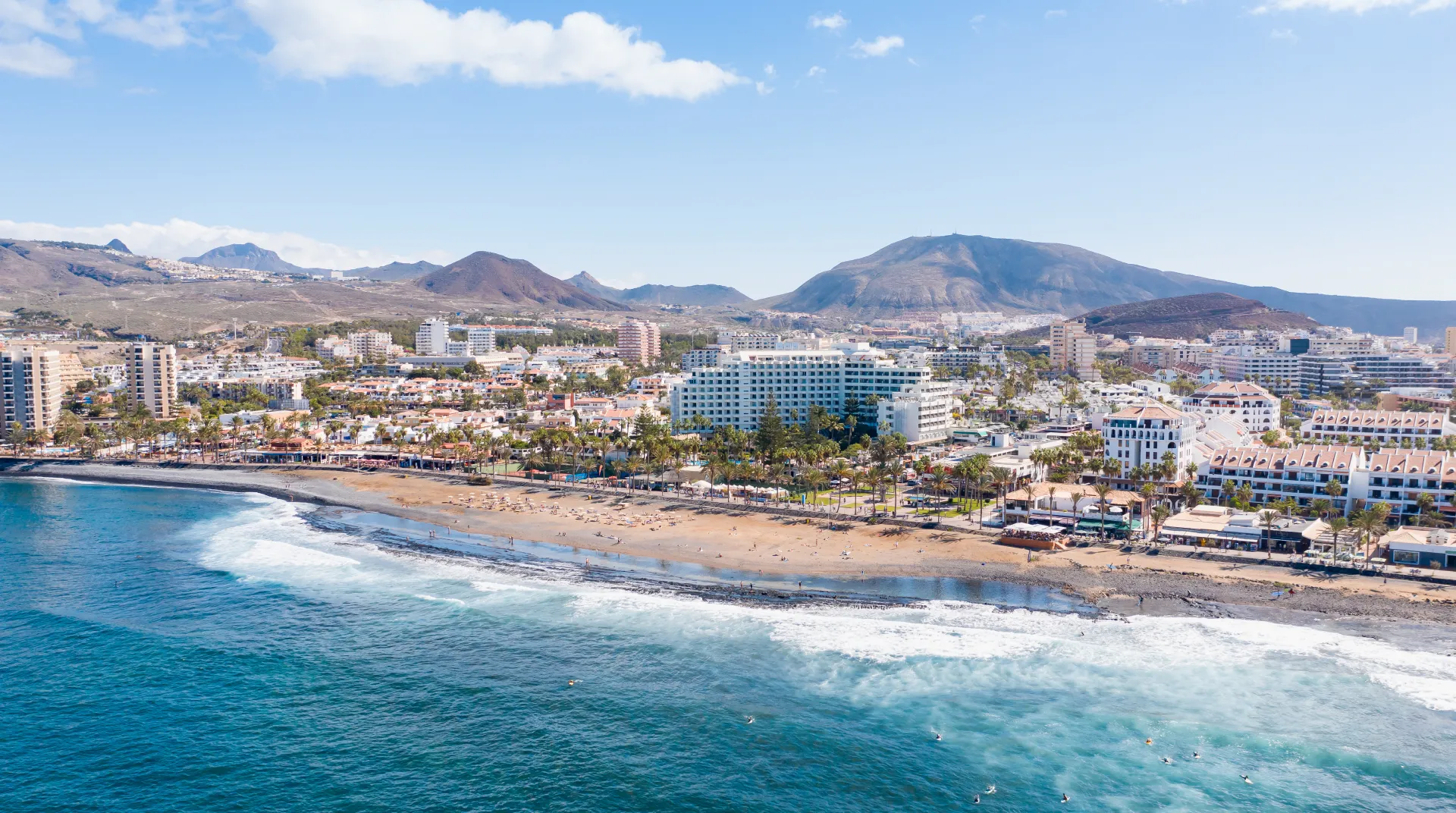 Playa con edificios, montañas al fondo y cielo despejado.