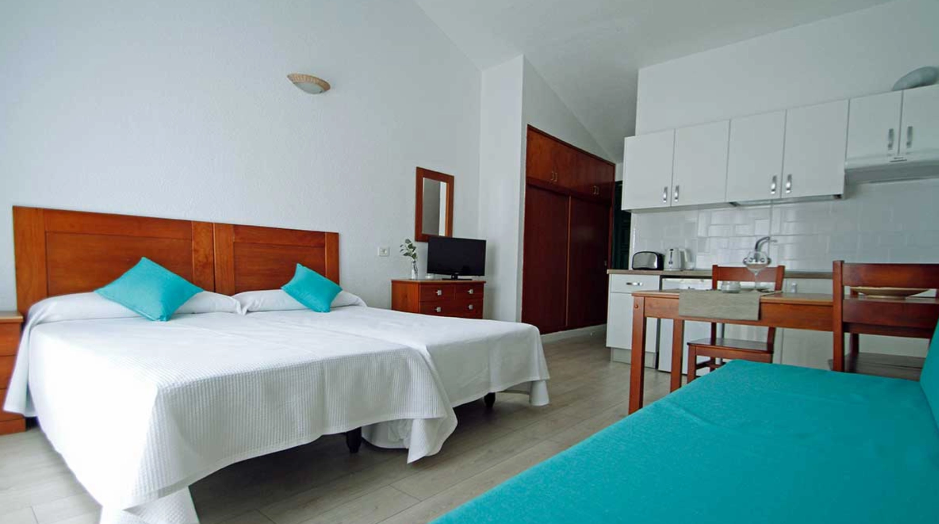 Habitación luminosa con cama doble, cocina pequeña, y decoración en blanco y madera.