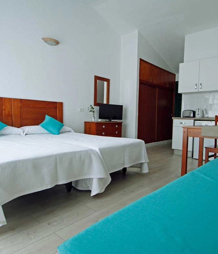 Habitación con dos camas, cojines azules, TV, tocador, cocina pequeña y sillas de madera.