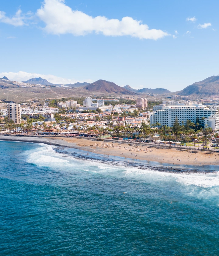Vista aérea de una playa con edificios y montañas al fondo, cielo azul y algunas nubes.
