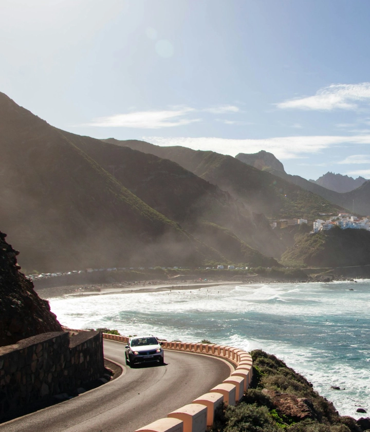 Carretera costera junto al mar y montañas, un coche circula bajo un cielo despejado.