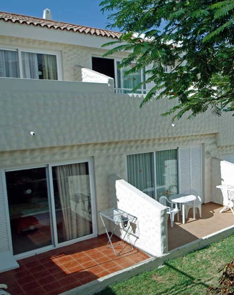 Casa de dos pisos blanca con balcón, terraza y muebles de exterior rodeada de césped y árboles.