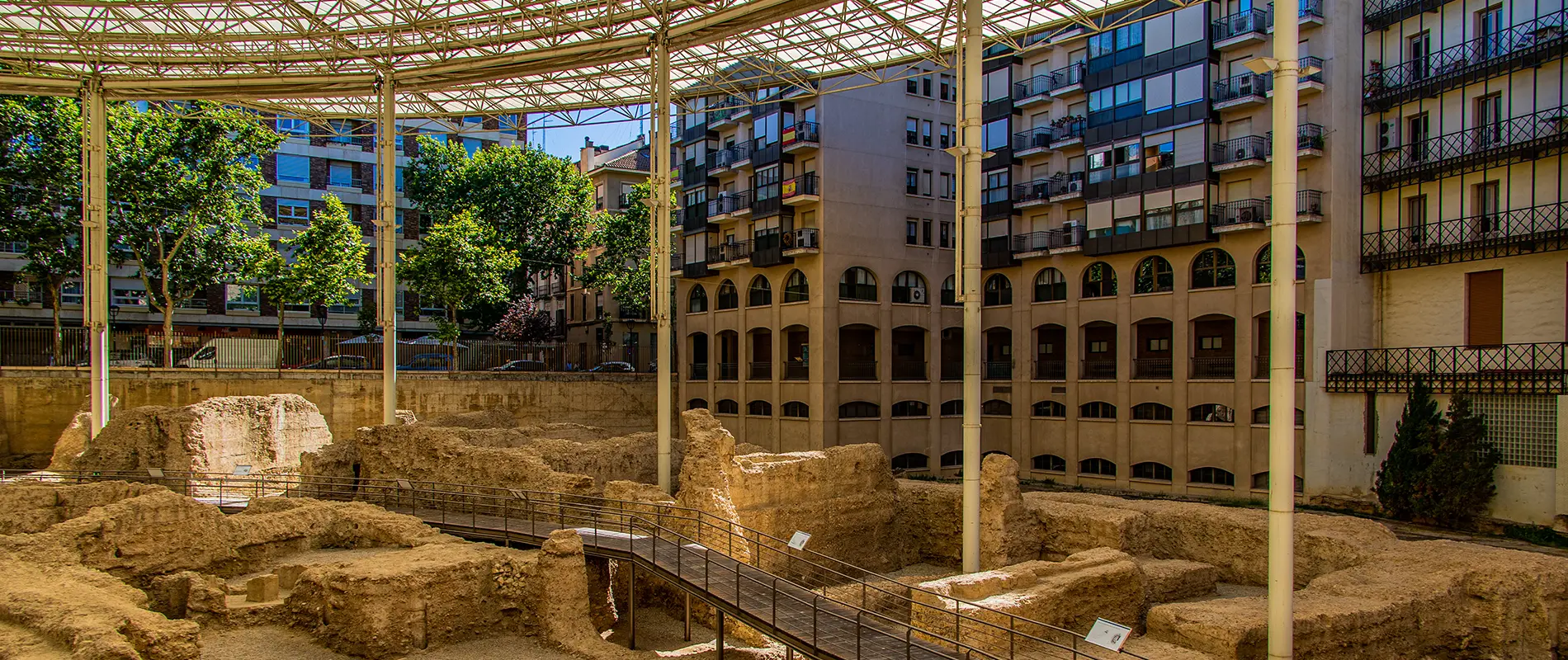 Ruinas arqueológicas cubiertas junto a edificios modernos y árboles en un área urbana.