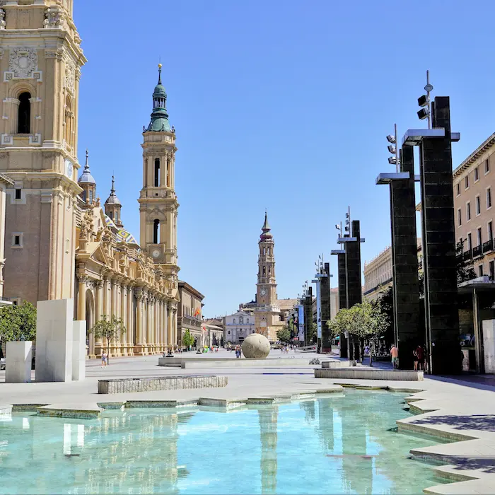 Plaza en Zaragoza con una iglesia, torre y fuente, bajo un cielo azul despejado.