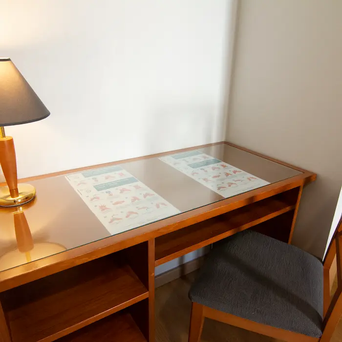 Escritorio de madera con vidrio, silla gris, lámpara y hojas debajo del cristal.