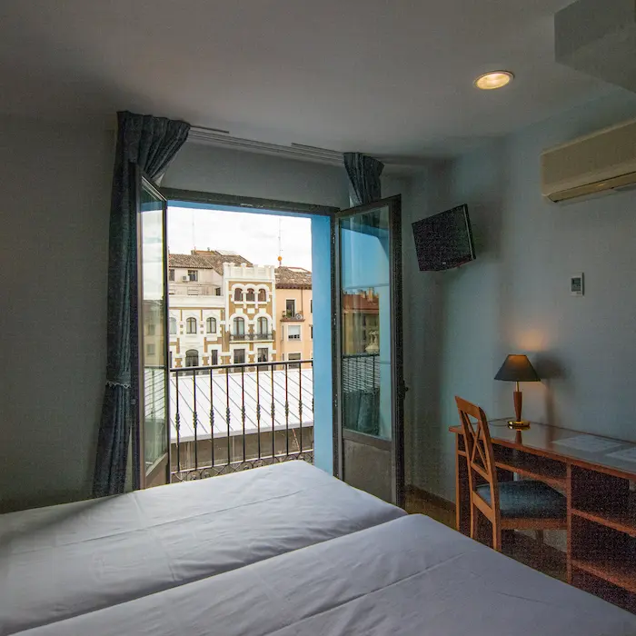 Habitación con cama doble, escritorio, lámpara y balcón con vista a edificios antiguos.