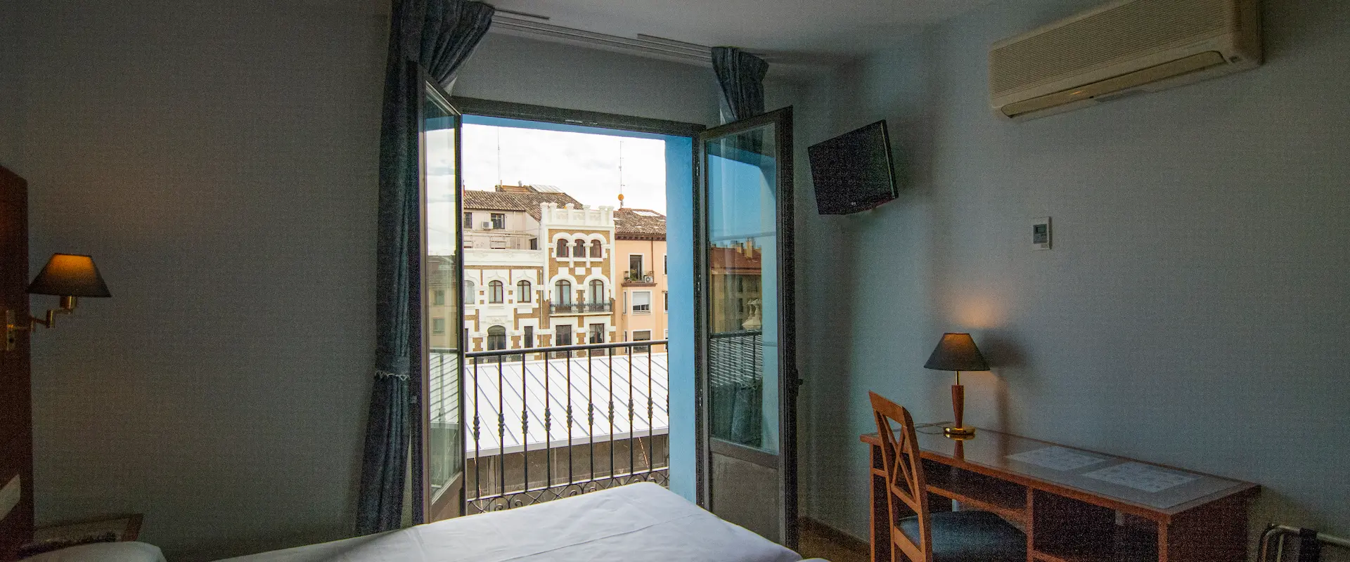 Habitación con balcón, vista a edificios, escritorio, lámpara y aire acondicionado en la pared.