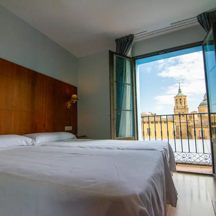 Habitación de hotel con dos camas, balcón y vista a una torre histórica bajo el cielo azul.