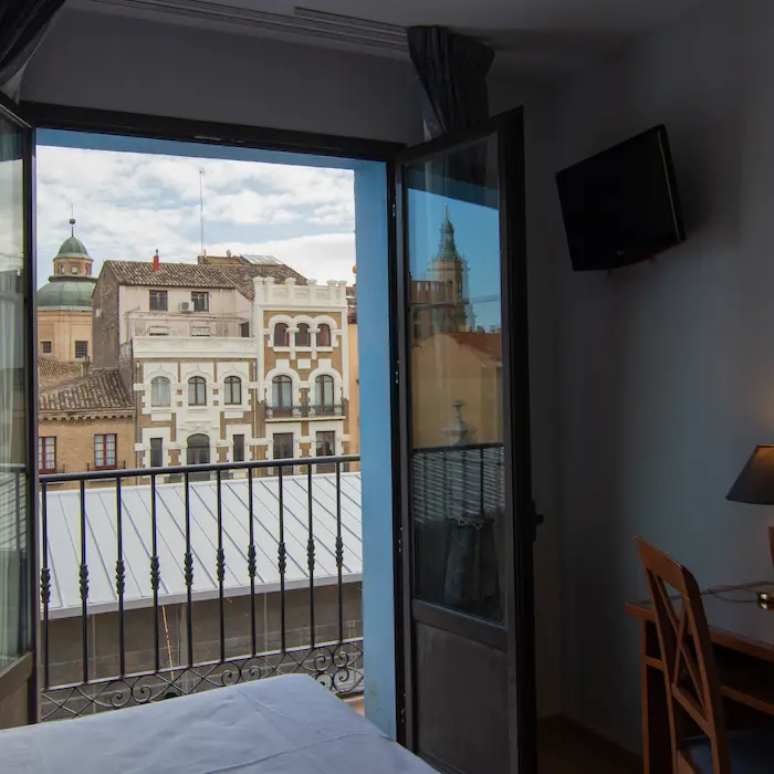 Vista desde una habitación con balcón hacia edificios antiguos y cielo parcialmente nublado.