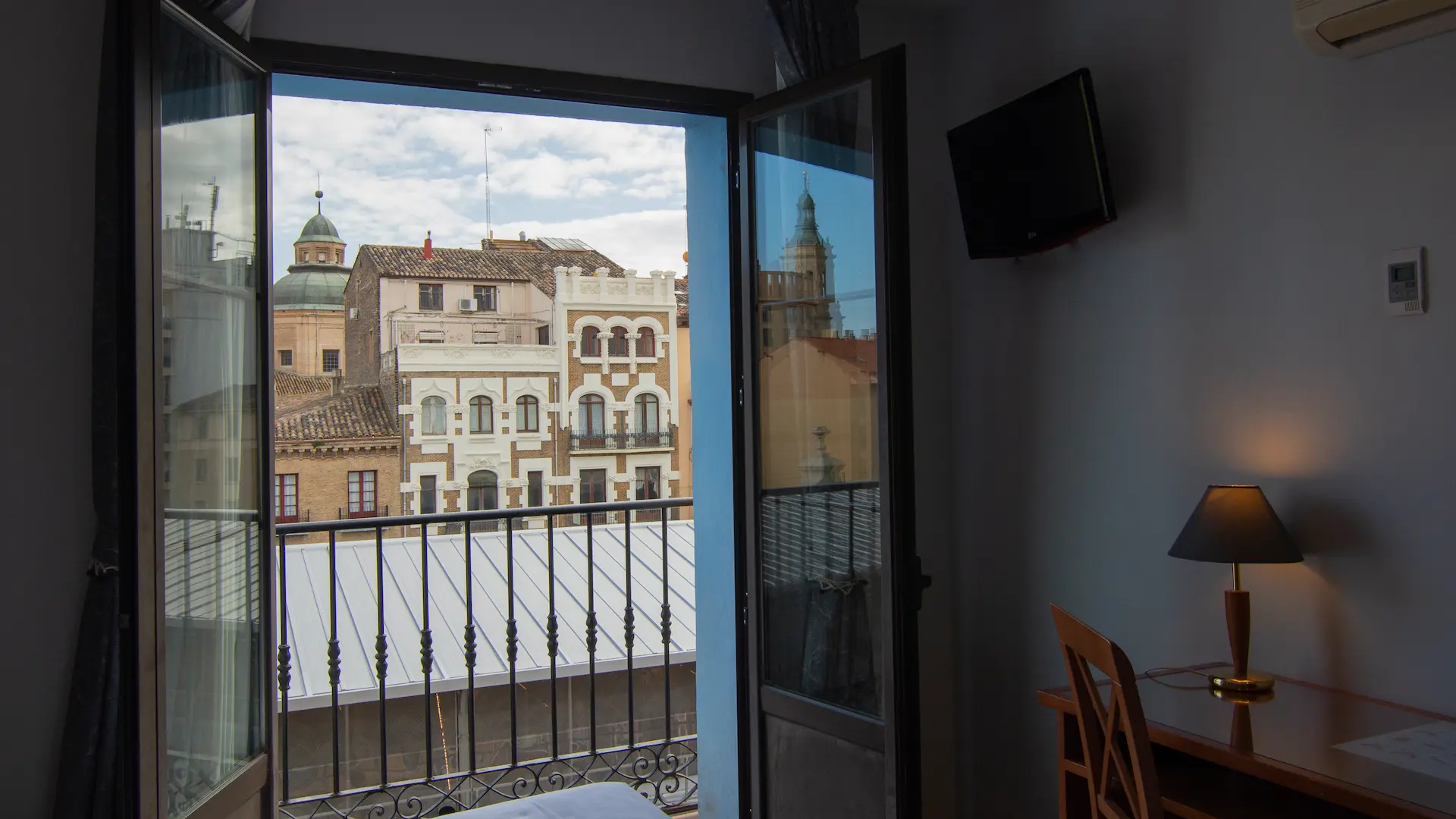 Habitación con vistas a edificios antiguos desde un balcón; lámpara encendida en el escritorio.