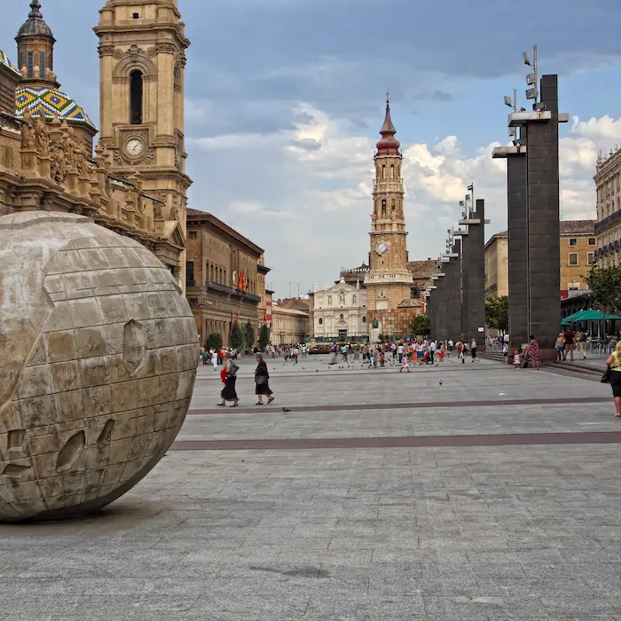 Plaza con una antigua catedral, una esfera de piedra y personas caminando bajo un cielo nublado.