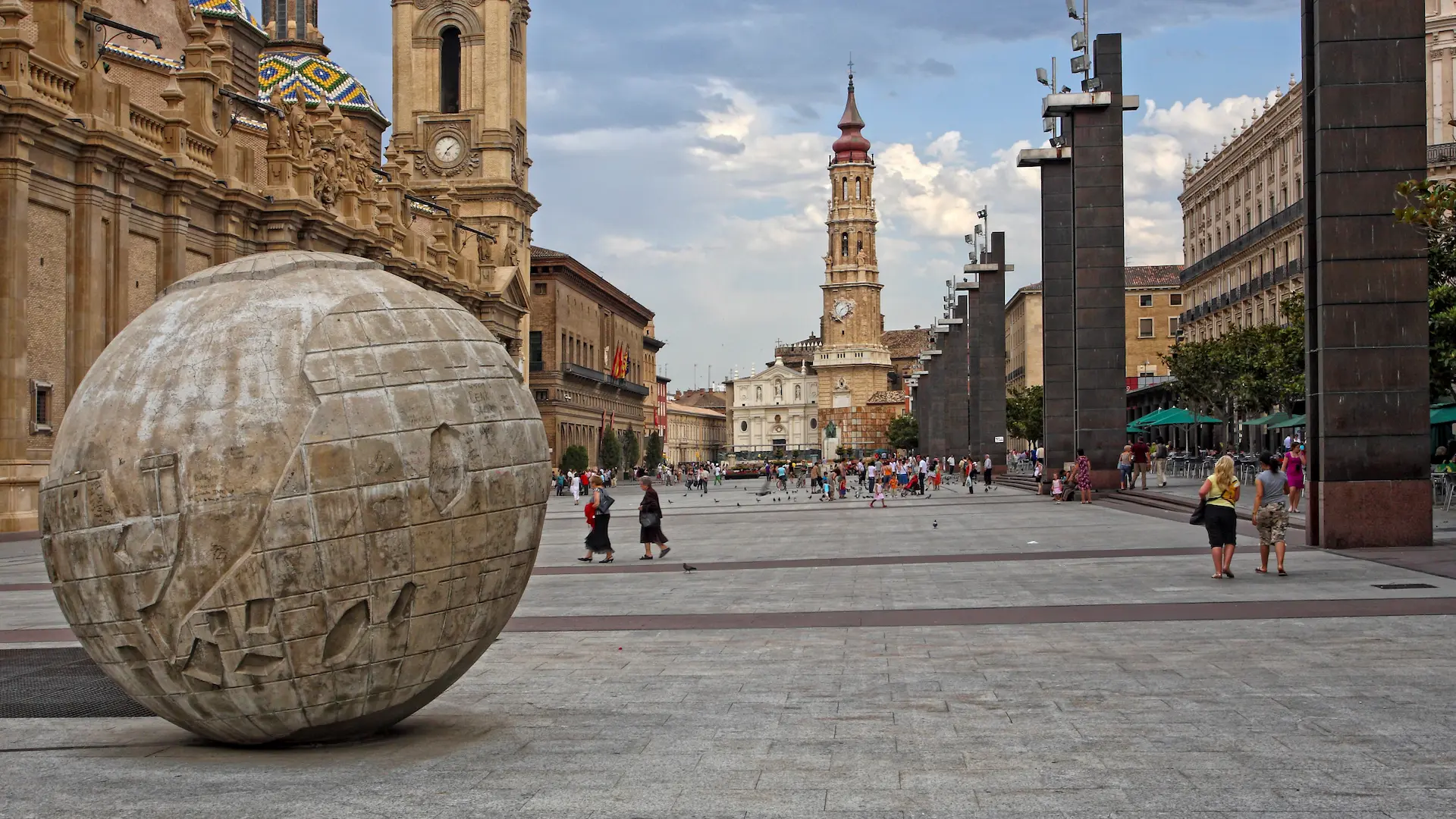 Plaza con escultura esférica y personas paseando cerca de edificios históricos y una torre.
