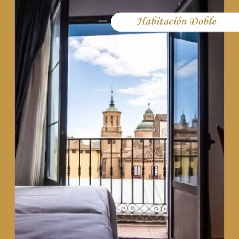 Habitación doble con balcón y vista a edificios históricos bajo un cielo azul con pocas nubes.